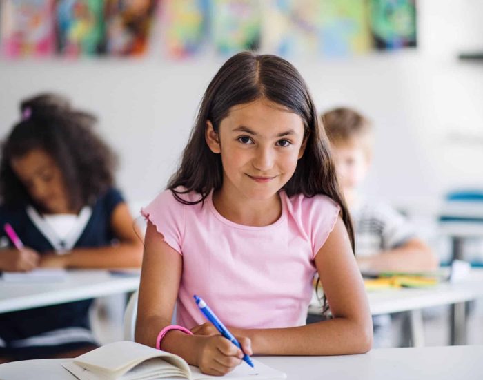 A portrait of small happy school girl sitting at the desk in classroom, looking at camera.