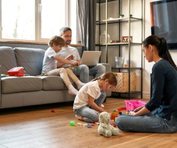 Young Caucasian family with children relax rest at home on quarantine or lockdown. Mother play with daughter toys, father work freelance online on computer. Parents and kids weekend indoors.