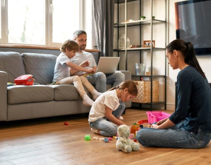 Young Caucasian family with children relax rest at home on quarantine or lockdown. Mother play with daughter toys, father work freelance online on computer. Parents and kids weekend indoors.